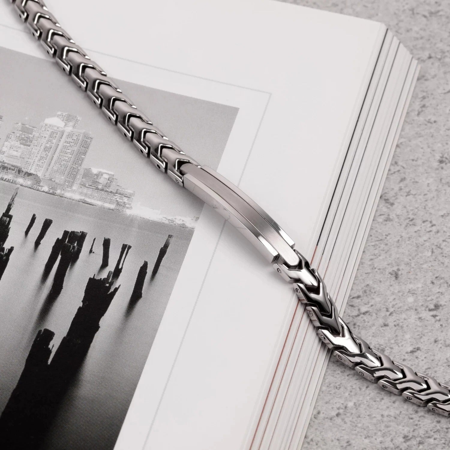 Aegon stainless steel bracelet styled across an open black-and-white photography book, showcasing its polished finish and V-link detail on a textured stone surface.