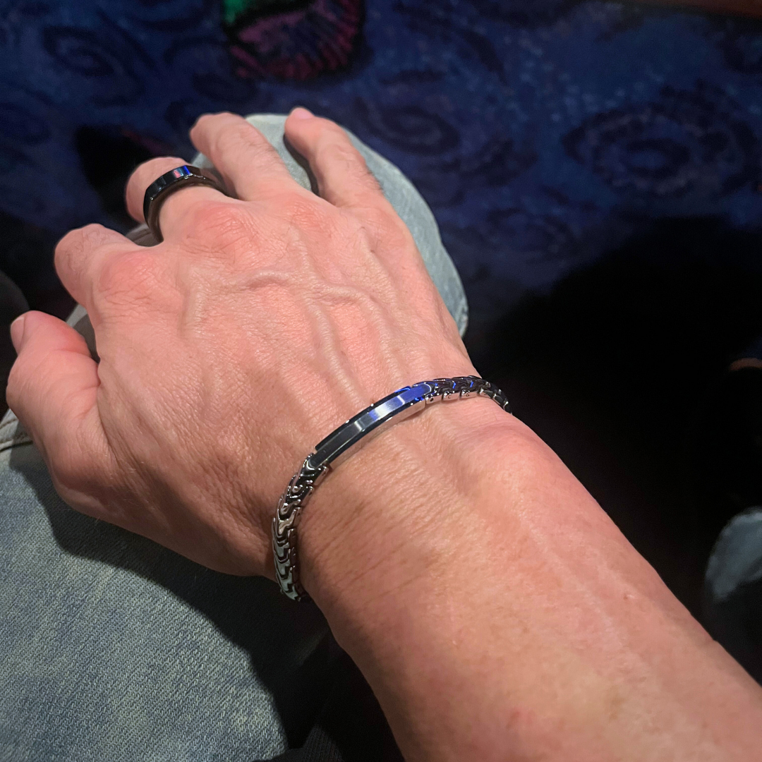 Close-up of a man's hand wearing the Aegon bracelet and a matching ring, with a faint blue light reflecting off the polished centerpiece of the bracelet.