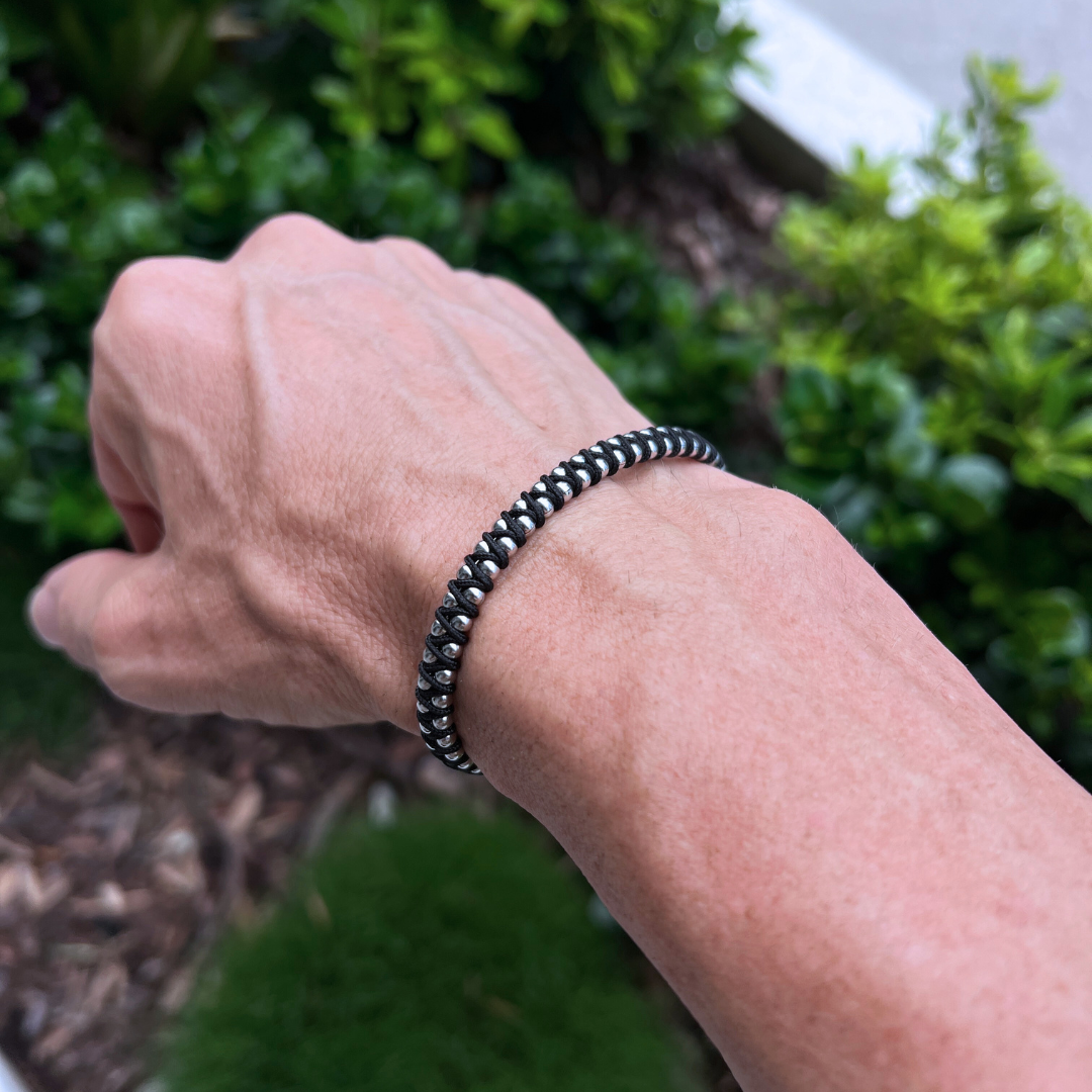 Outdoor wrist shot of the Armoured Elegance Synergy black bracelet, featuring stainless steel beads woven with black cord for a modern masculine look.