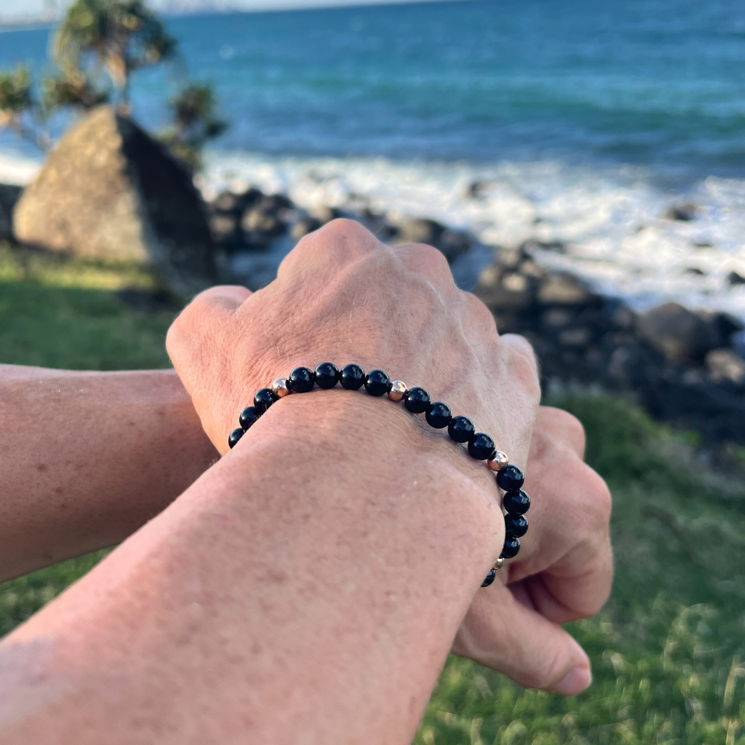 Auric Lineage Shadow edition black bead bracelet worn near the coastline with ocean waves behind.
