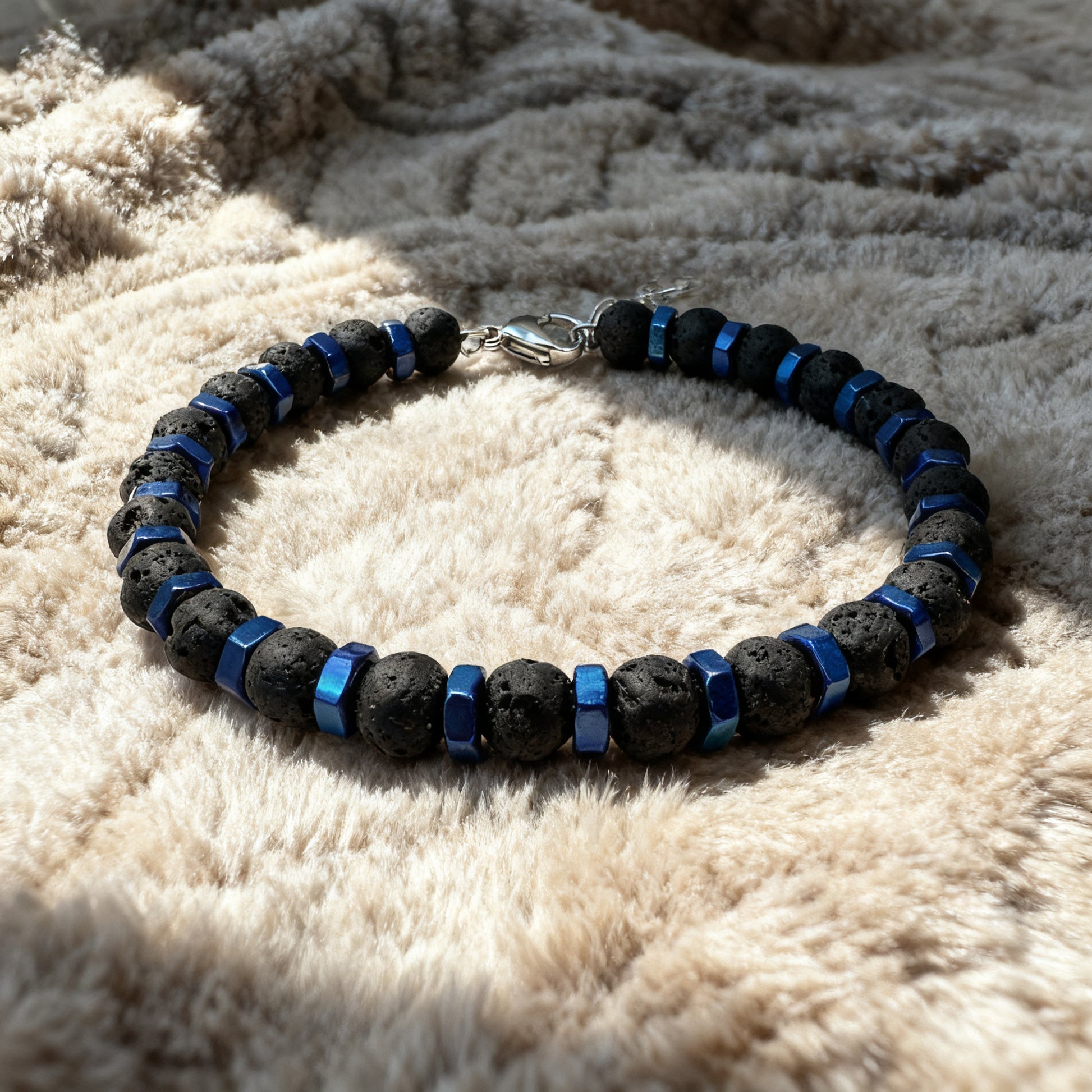 Luxury men’s Blue Nexus bracelet featuring black lava stones and anodized blue hex nut spacers with stainless-steel clasp, displayed on soft beige fabric.
