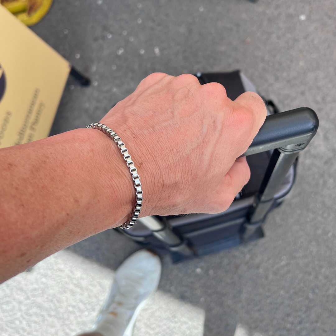 Element stainless steel box-chain bracelet worn while pulling a suitcase handle in a travel setting.
