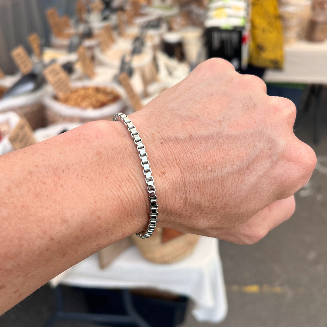 Element stainless steel box-chain bracelet worn on wrist in an outdoor market lifestyle setting.