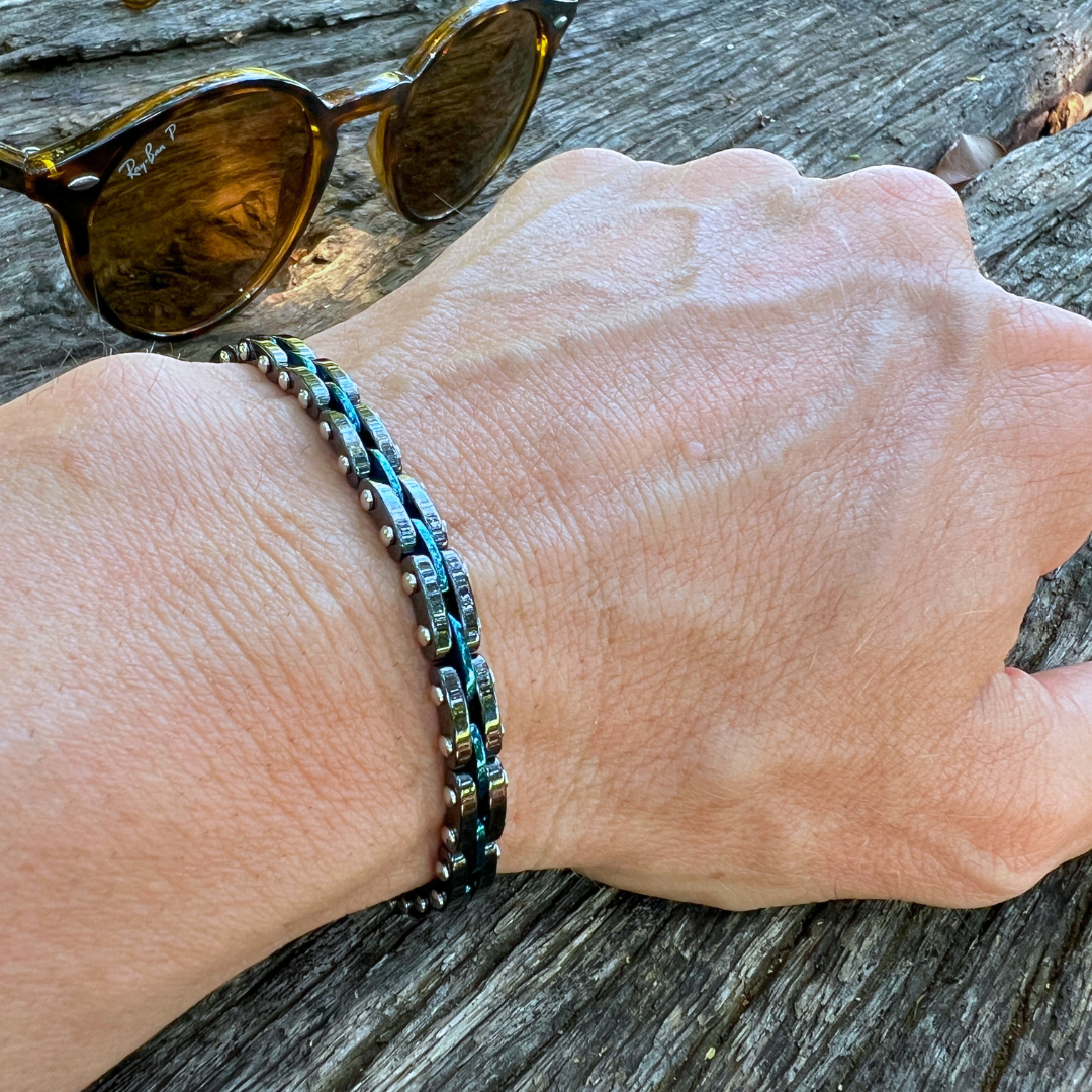 Close-up of the Velocity bracelet featuring blue and black stainless steel chain links, worn on a wrist outdoors with sunglasses in background.