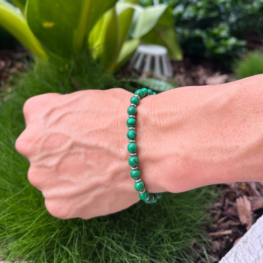 Close-up of Verdure malachite bracelet on wrist surrounded by greenery, showcasing polished green beads and stainless steel ringlets.