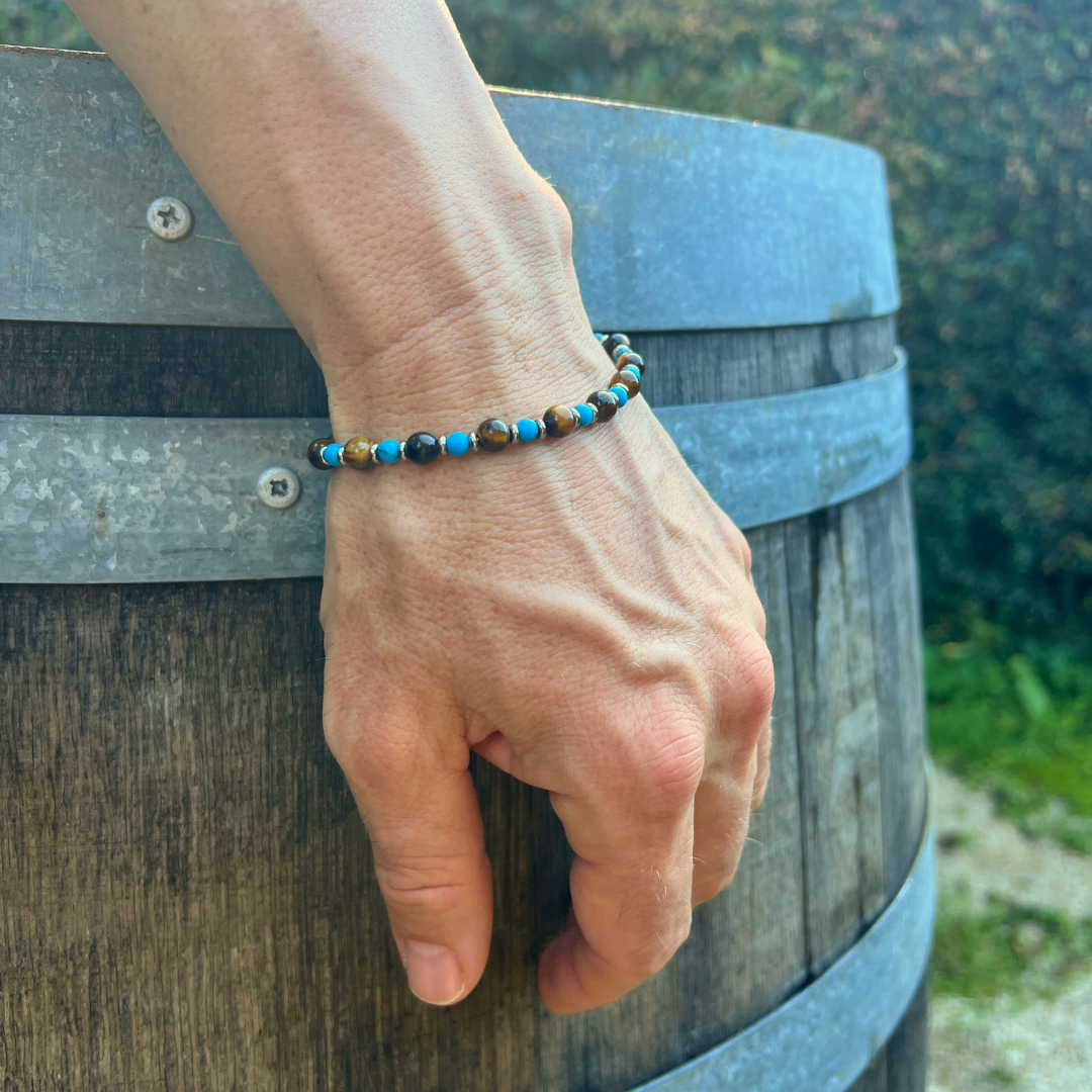 The Wild Oasis bead bracelet displayed on a man’s wrist resting against a rustic barrel, showing the full range of blue, brown, and black beads in natural light.